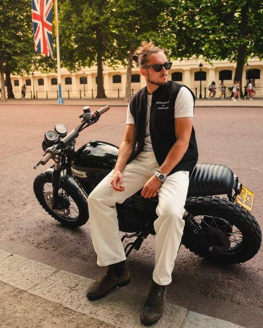 Man wearing The Canvas Vest by Scrambler of London, leaning on a Triumph Scrambler motorcycle with sunglasses and Union Jack flags in the background.