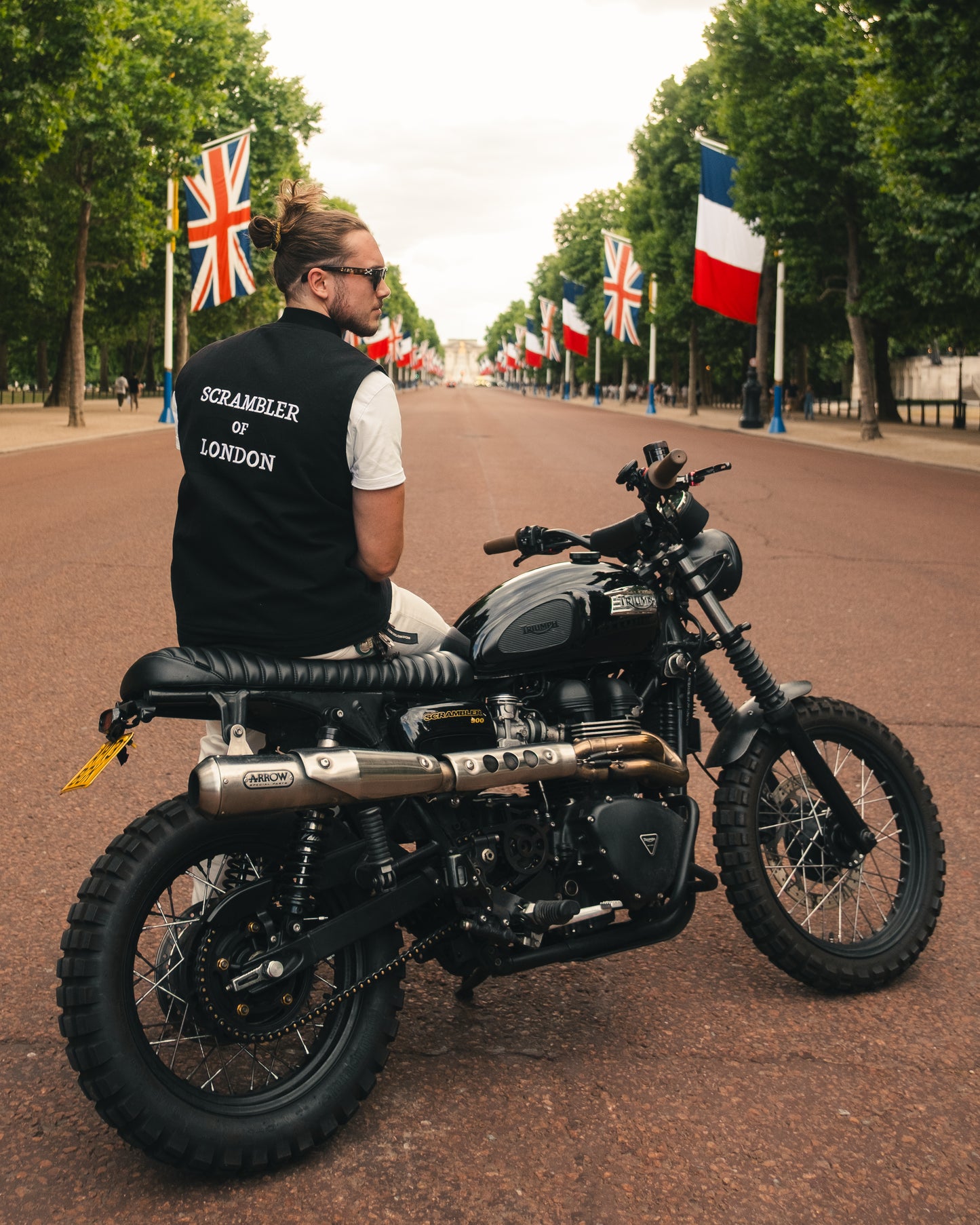 Man wearing The Canvas Vest by Scrambler of London, sitting on a Triumph Scrambler motorcycle with sunglasses and Union Jack flags in the background.