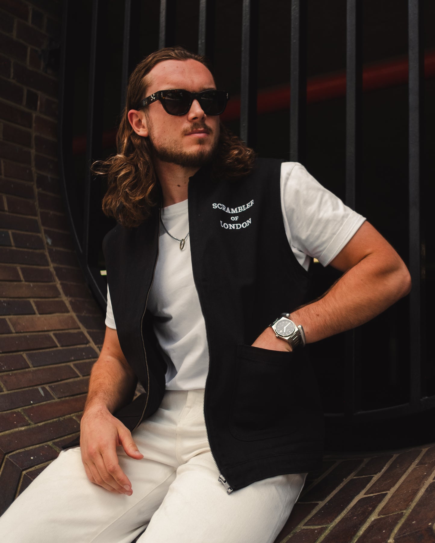 Man wearing The Canvas Vest by Scrambler of London, sitting on a wall in the sun with sunglasses and a smart watch on his wrist.
