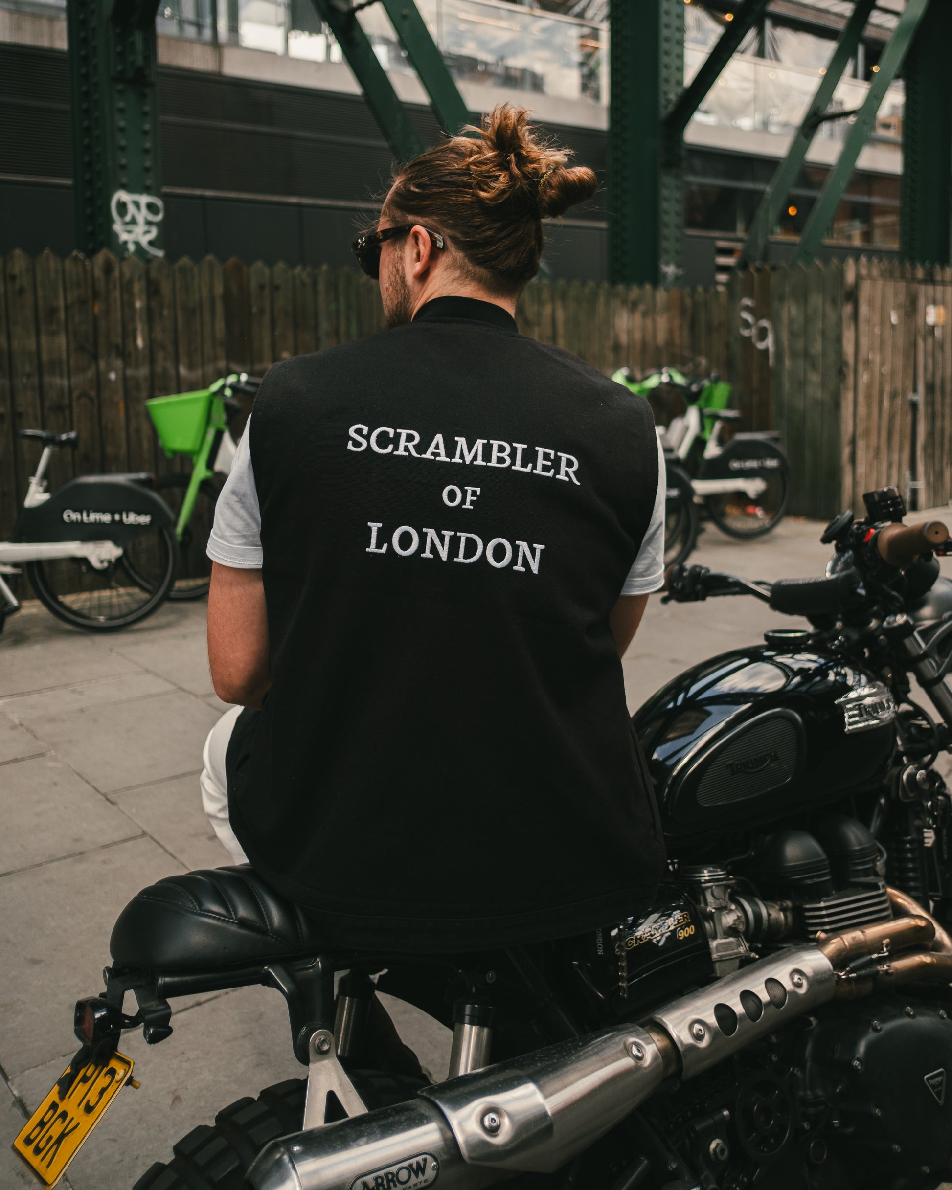 Man wearing The Canvas Vest by Scrambler of London, leaning on a Triumph Scrambler motorcycle with sunglasses and London in the background.