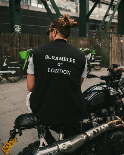Man wearing The Canvas Vest by Scrambler of London, leaning on a Triumph Scrambler motorcycle with sunglasses and London in the background.